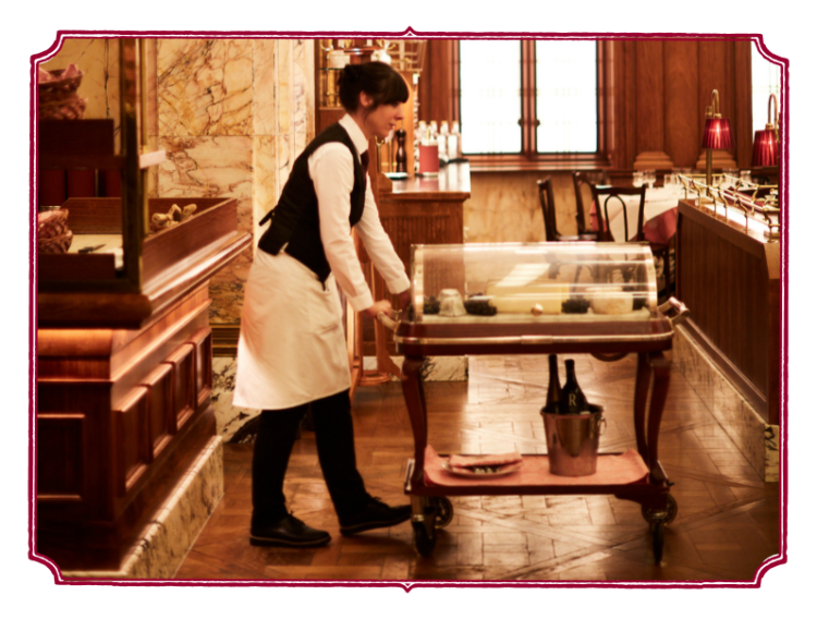 Waiter in a vintage-style restaurant pushing a dessert or cheese cart with a glass cover, set in a richly decorated interior with wooden and marble accents.